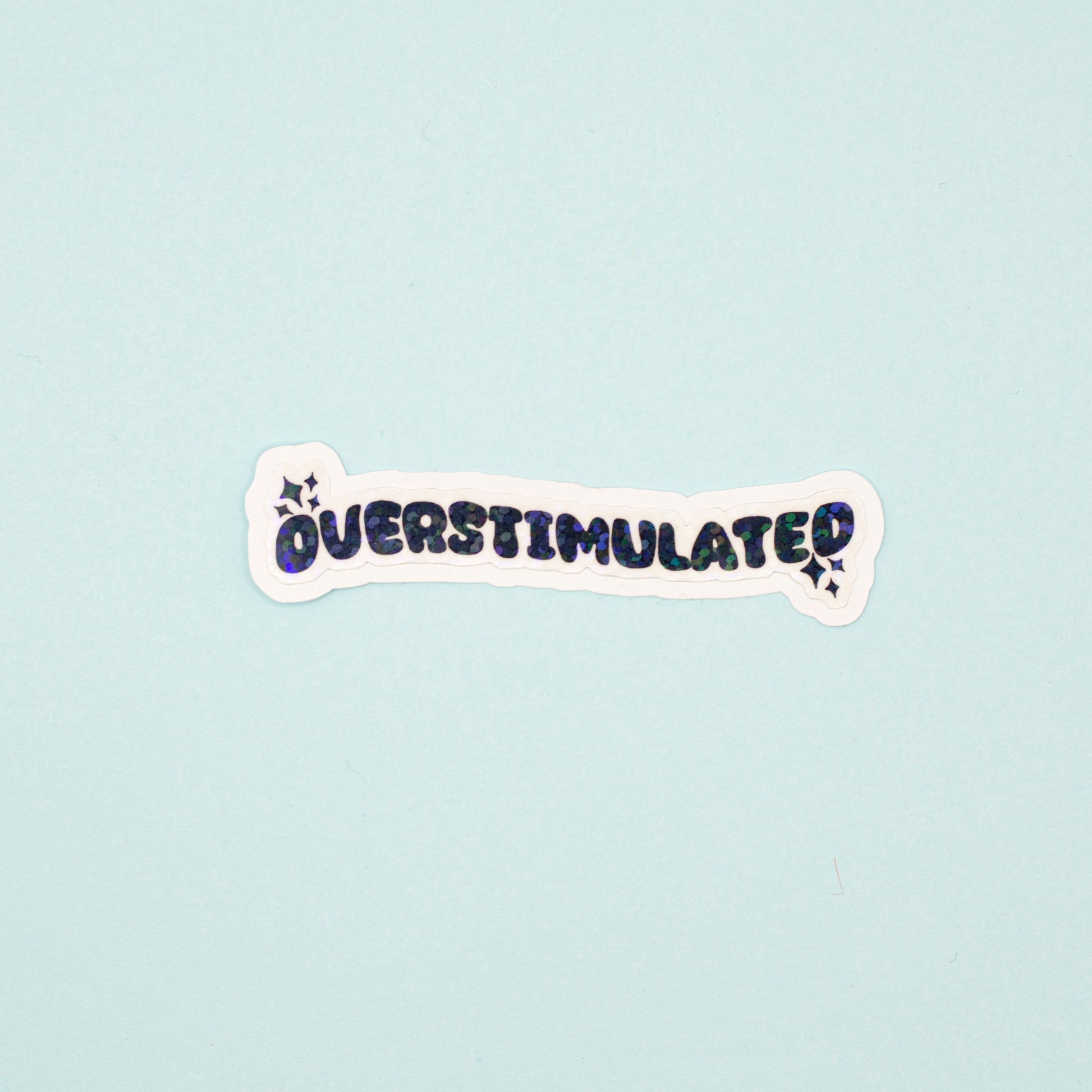 A die-cut sticker with the word 'OVERSTIMULATED' in black bubble letters and a holographic dot overlay on a white background,  shown on a light blue background.