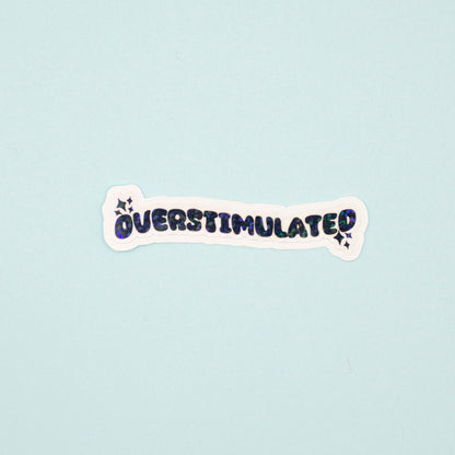 A die-cut sticker with the word 'OVERSTIMULATED' in black bubble letters and a holographic dot overlay on a white background,  shown on a light blue background.