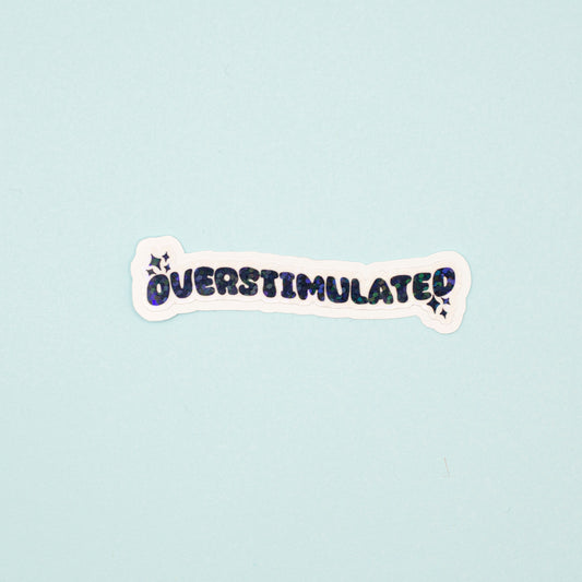 A die-cut sticker with the word 'OVERSTIMULATED' in black bubble letters and a holographic dot overlay on a white background,  shown on a light blue background.