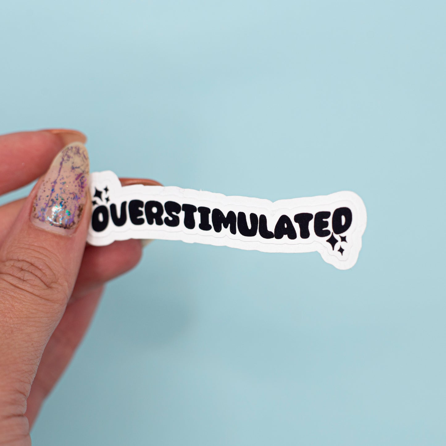 a white die-cut sticker that reads 'OVERSTIMULATED' in bold black letters against a light blue background.