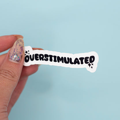 a white die-cut sticker that reads 'OVERSTIMULATED' in bold black letters against a light blue background.