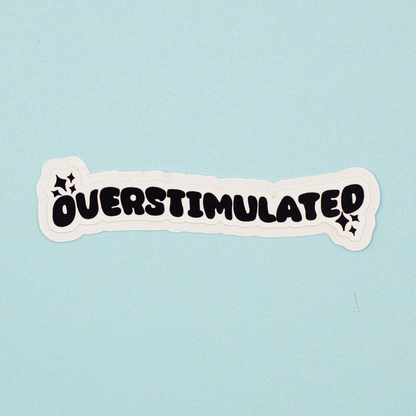 A die-cut sticker with the word 'OVERSTIMULATED' in bold black bubble letters on a white background, shown on a light blue surface.