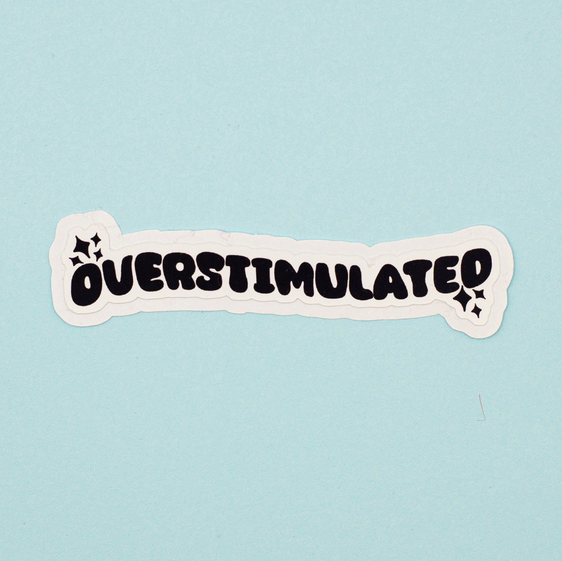 A die-cut sticker with the word 'OVERSTIMULATED' in bold black bubble letters on a white background, shown on a light blue surface.