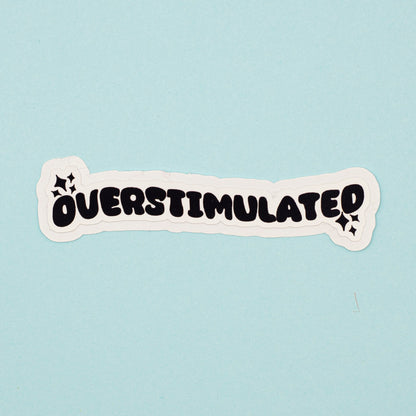 A die-cut sticker with the word 'OVERSTIMULATED' in bold black bubble letters on a white background, shown on a light blue surface.