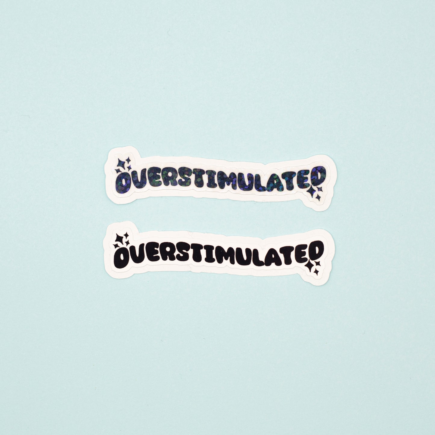 Two die-cut stickers with the word 'OVERSTIMULATED' in a black bubbly font pne matte, one with a holographic dot overlay, displayed on a light blue background.
