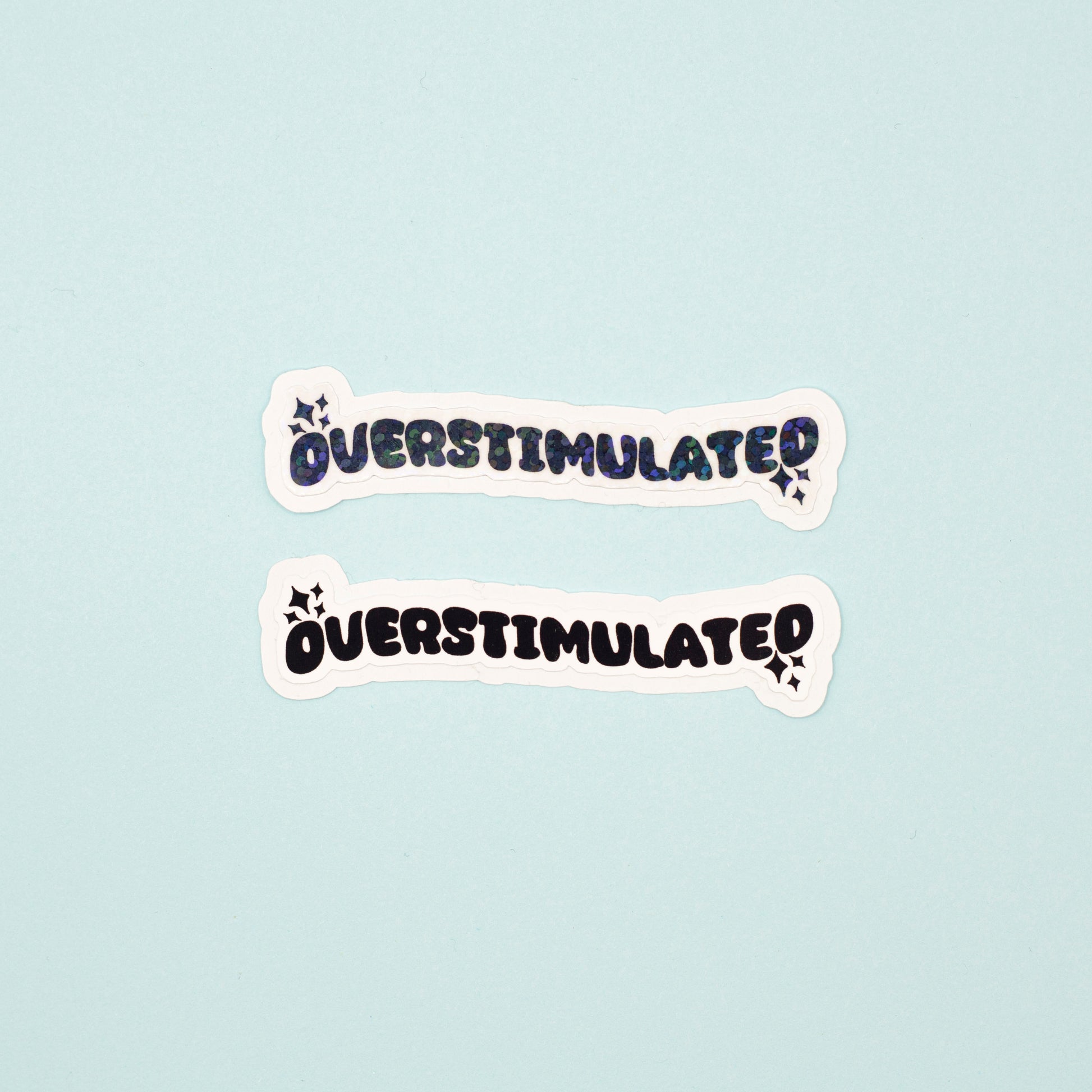 Two die-cut stickers with the word 'OVERSTIMULATED' in a black bubbly font pne matte, one with a holographic dot overlay, displayed on a light blue background.