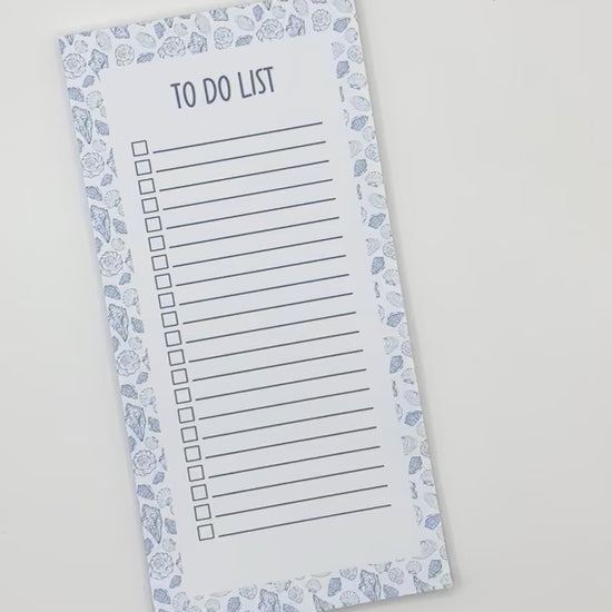A vertical to-do list notepad with a blue and white sea shell border, displayed diagonally on a light off-white background.