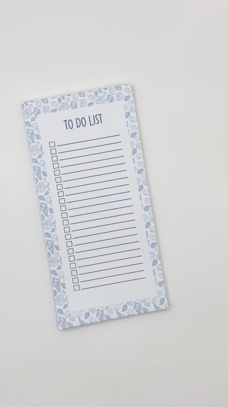 A vertical to-do list notepad with a blue and white sea shell border, displayed diagonally on a light off-white background.