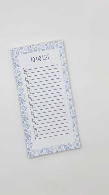 A vertical to-do list notepad with a blue and white sea shell border, displayed diagonally on a light off-white background.