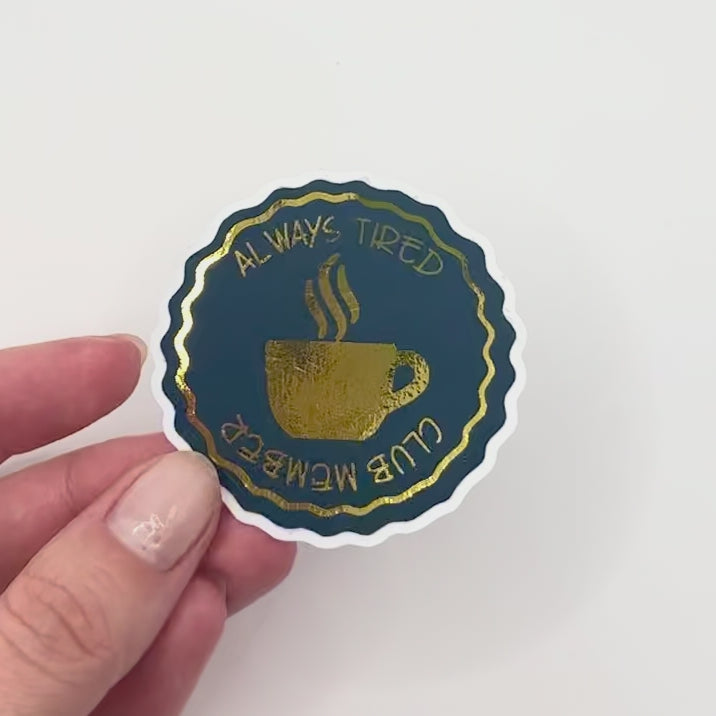 Circular sticker with a coffee cup design and text 'Always Tired Club Member' with gold details on white background. 