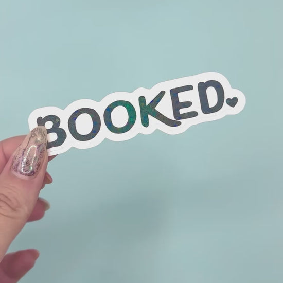 a die-cut sticker that reads 'BOOKED.' in black letters with a holographic overlay with a white outline, set against a solid light blue background.