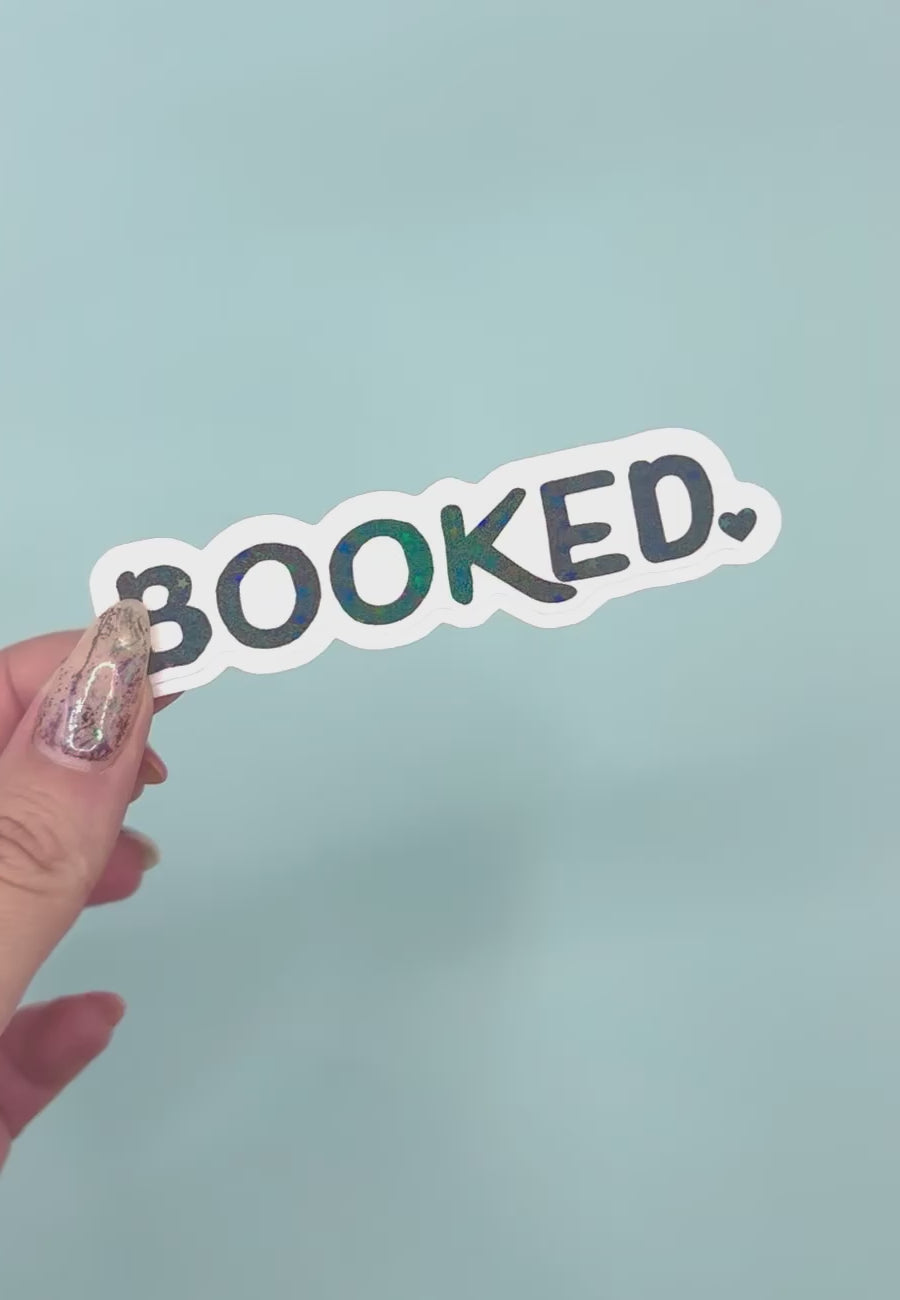 a die-cut sticker that reads 'BOOKED.' in black letters with a holographic overlay with a white outline, set against a solid light blue background.