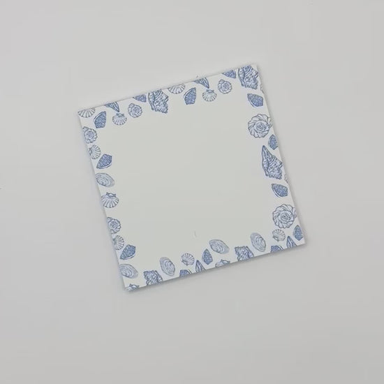 Square memo pad with a blue and white sea shell border, displayed diagonally on a light off-white background.