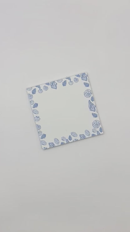 Square memo pad with a blue and white sea shell border, displayed diagonally on a light off-white background.