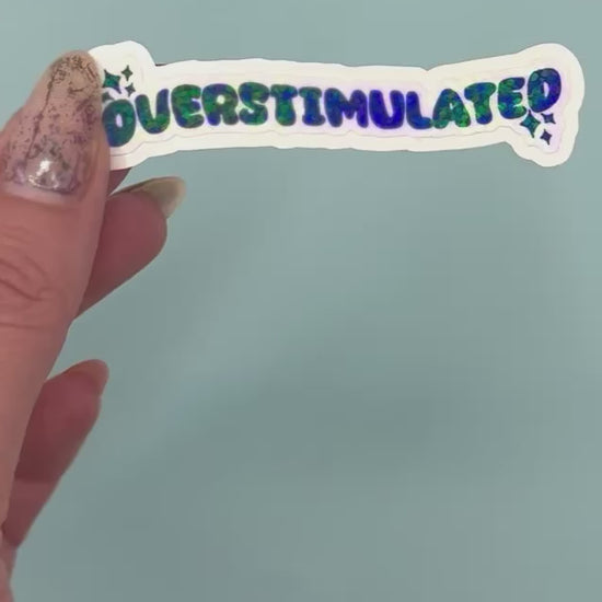 A die-cut sticker with the word 'OVERSTIMULATED' in black bubble letters and a holographic dot overlay on a white background,  shown on a light blue background.