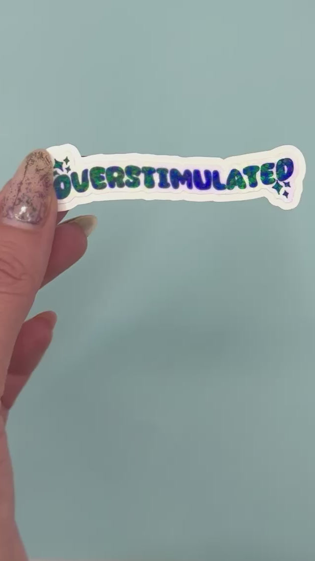A die-cut sticker with the word 'OVERSTIMULATED' in black bubble letters and a holographic dot overlay on a white background,  shown on a light blue background.