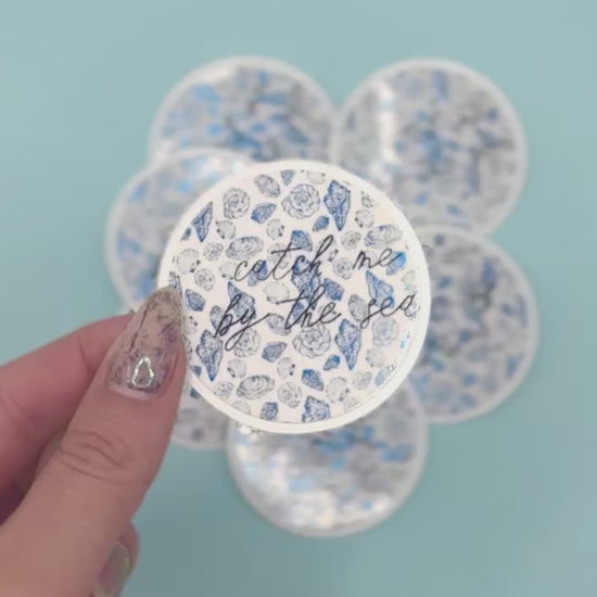  a round sticker that reads 'catch me by the sea' in black script over a metalic blue seashell pattern. More round stickers are blurred in the background against a light blue backdrop.