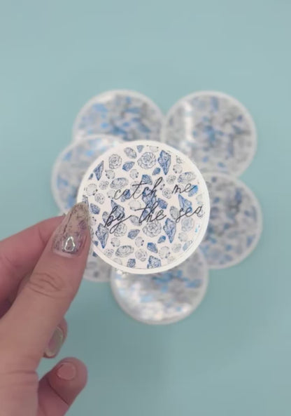  a round sticker that reads 'catch me by the sea' in black script over a metalic blue seashell pattern. More round stickers are blurred in the background against a light blue backdrop.