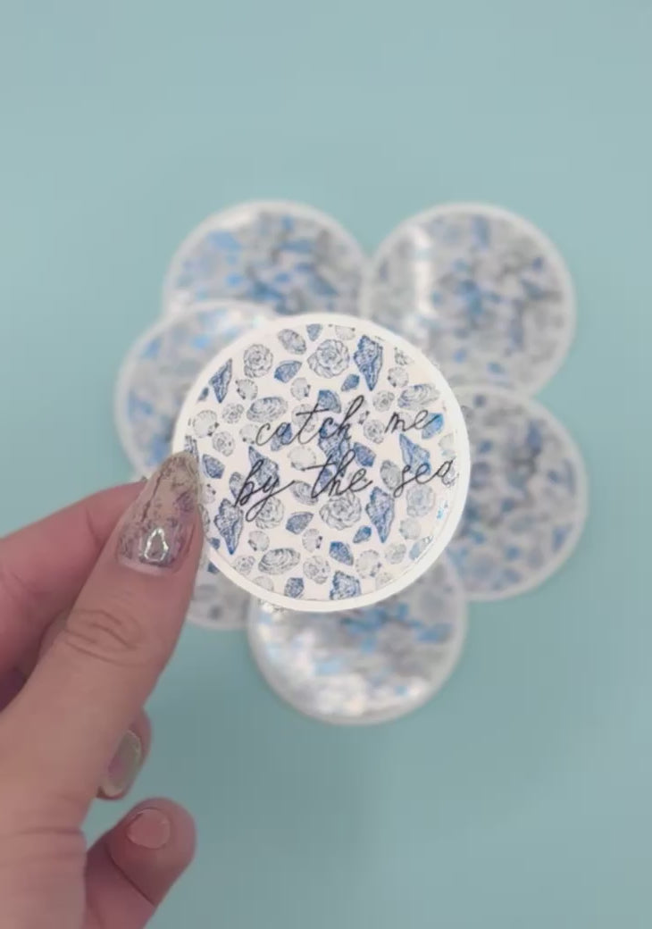  a round sticker that reads 'catch me by the sea' in black script over a metalic blue seashell pattern. More round stickers are blurred in the background against a light blue backdrop.