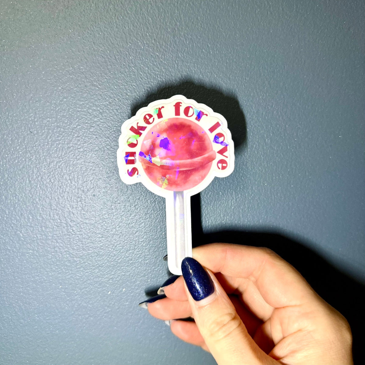 Hand holding a lollipop-shaped sticker with 'sucker for love' text and holographic finish against a blue background