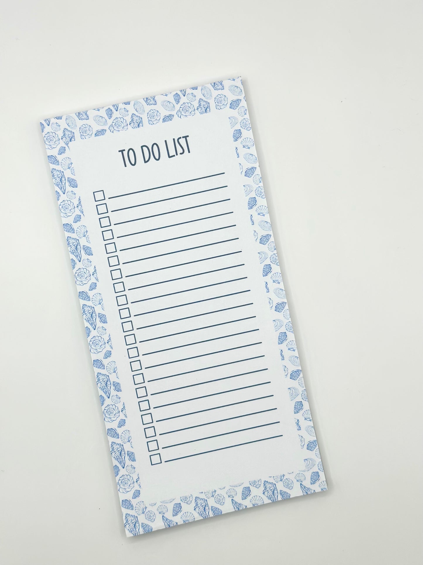 A vertical to-do list notepad with a blue and white sea shell border, displayed diagonally on a light off-white background.