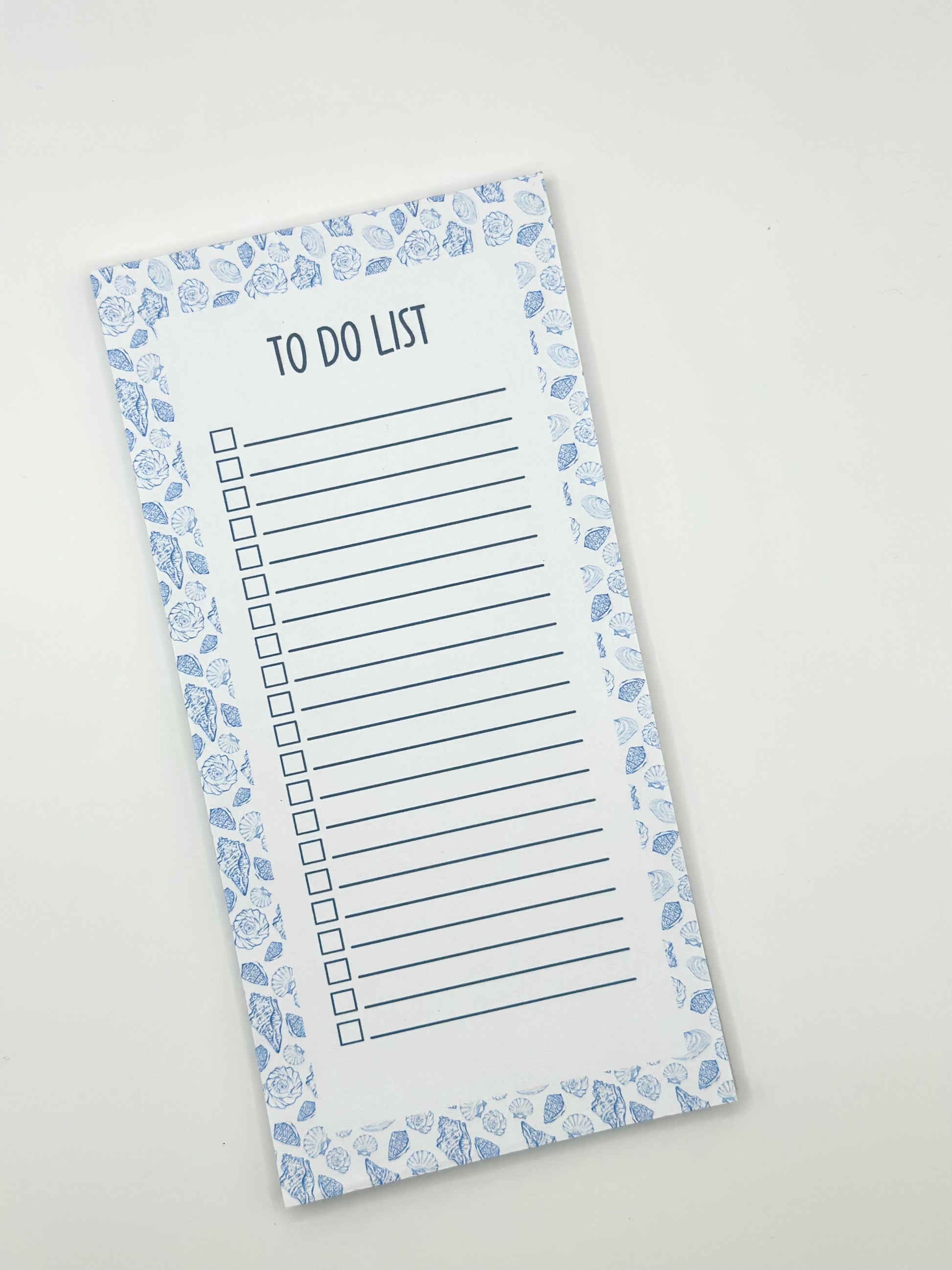 A vertical to-do list notepad with a blue and white sea shell border, displayed diagonally on a light off-white background.