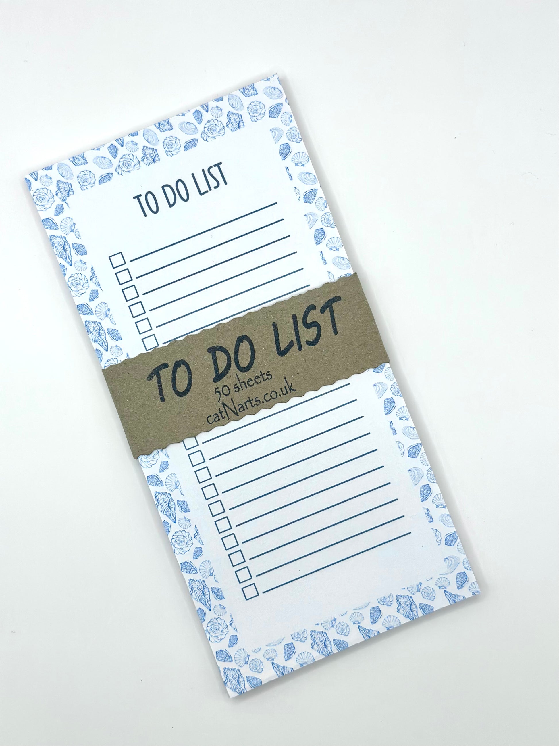 A vertical to-do list notepad with a blue and white sea shell border, displayed diagonally on a light off-white background with a craft paper band around.