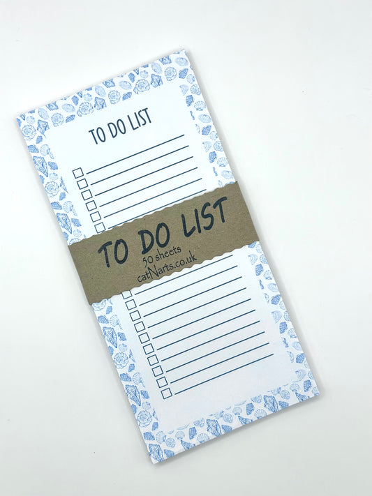 A vertical to-do list notepad with a blue and white sea shell border, displayed diagonally on a light off-white background with a craft paper band around.